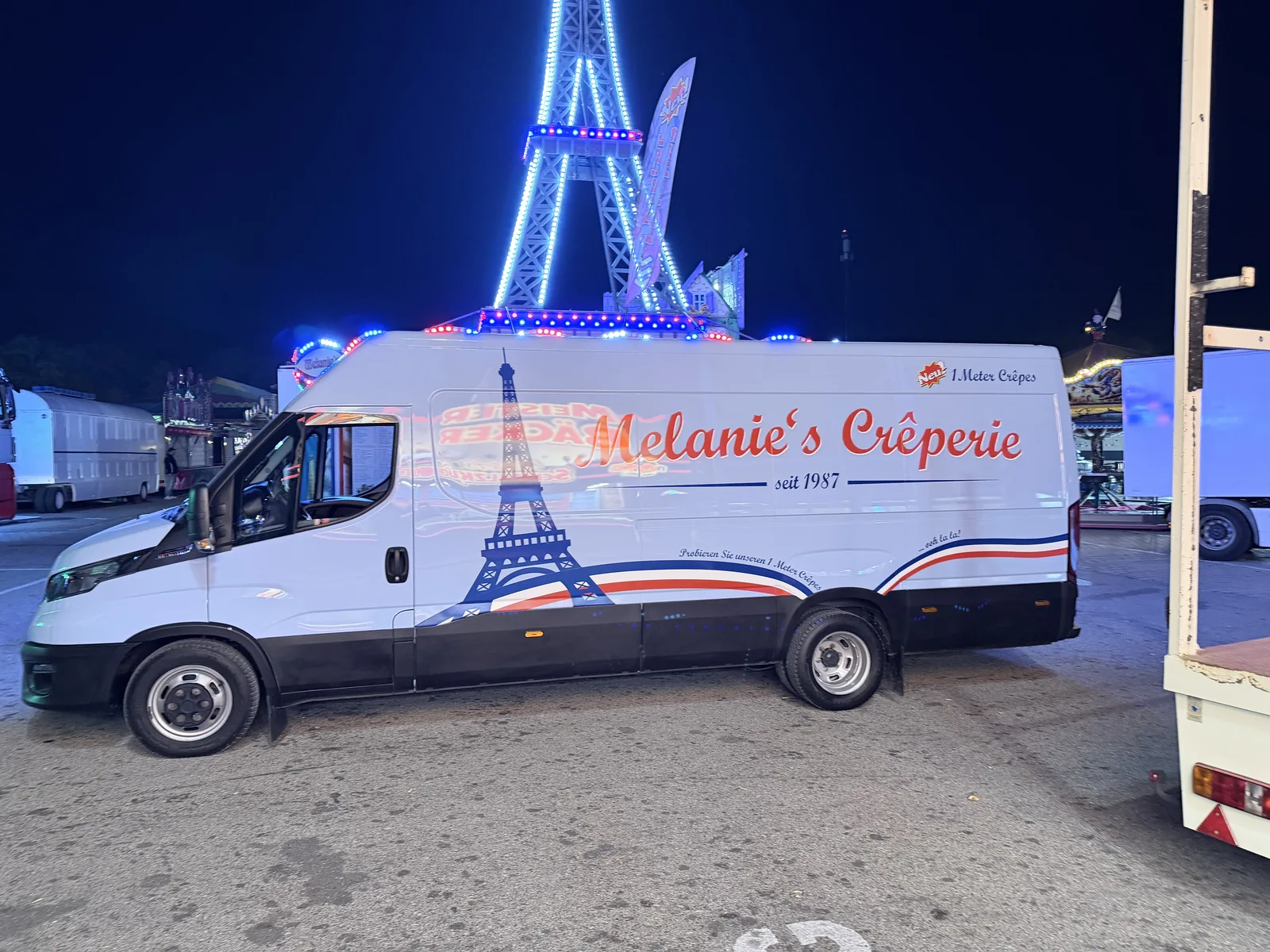 Der Bus mit dem Aufdruck „Melanie's Crêperie“ und einer Eiffelturm-Grafik, dahinter der leuchtende Turm.