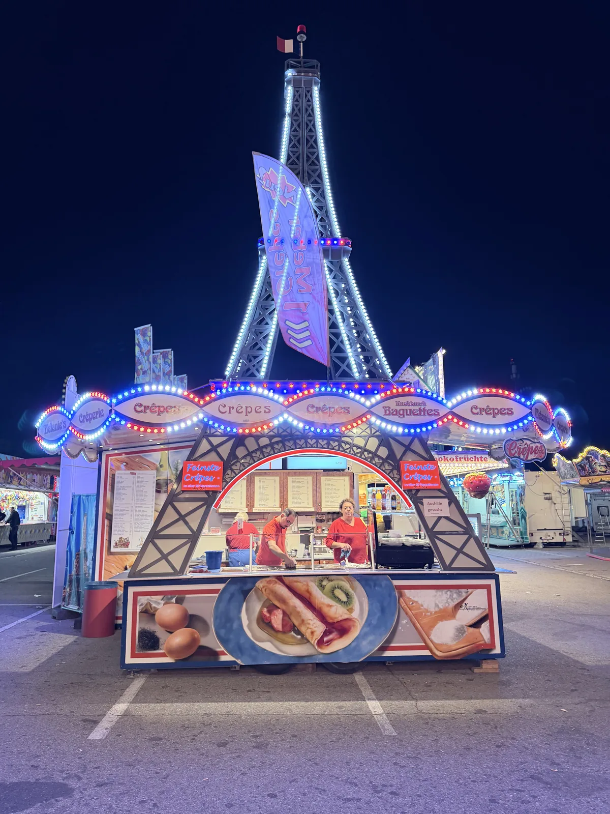 Der Stand bei Nacht von vorn: der Turm leuchtet weiß, die Bänder und Bogenschilder in Rot und Blau.